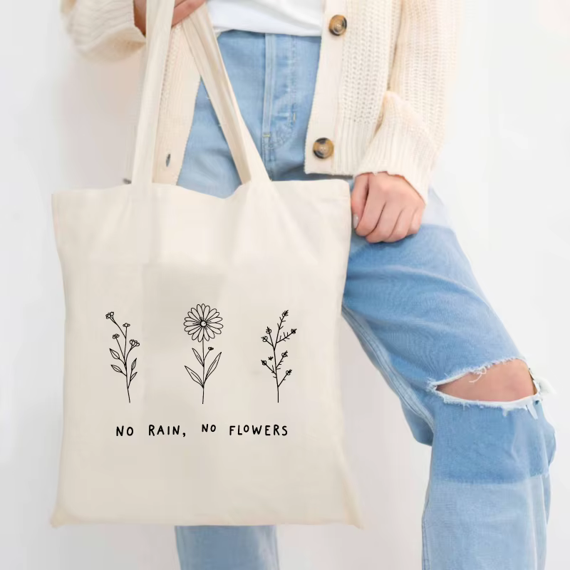 Sac fourre-tout en coton à motif floral crème et texte "No Rain No Flower", adapté à un usage quotidien et au shopping.