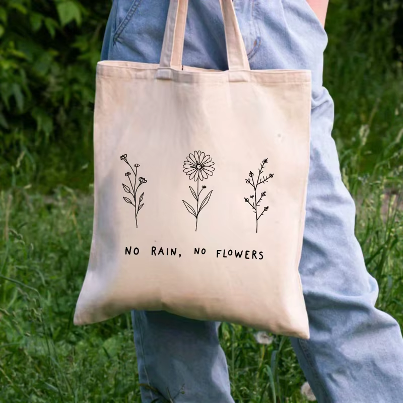 Sac fourre-tout en coton à motif floral crème et texte "No Rain No Flower", adapté à un usage quotidien et au shopping.